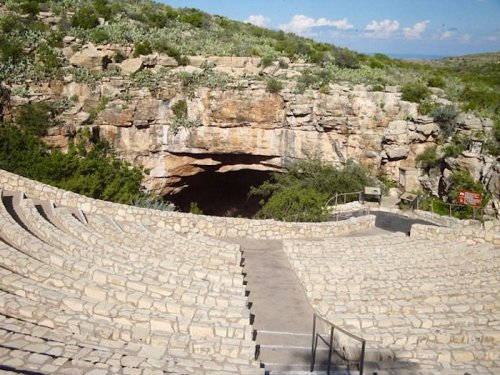 An outdoor amphitheater with stone seating, overlooking a cave entrance surrounded by lush greenery and blue skies.