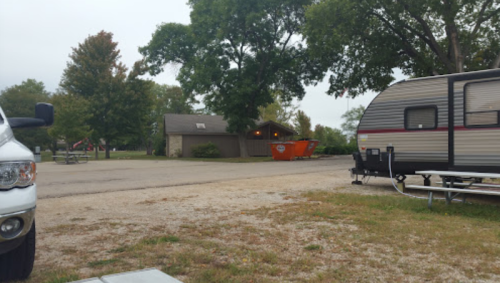 A campsite scene featuring a parked RV, a dumpster, and a building surrounded by trees on a cloudy day.
