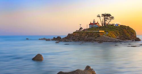 A serene coastal scene featuring a lighthouse on a rocky island at sunset, with calm waters and soft pastel skies.