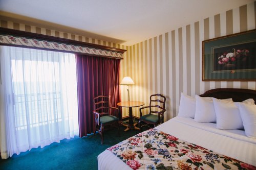 Cozy hotel room featuring a bed, two chairs, a table, and large windows with curtains. Floral decor on the bedspread.