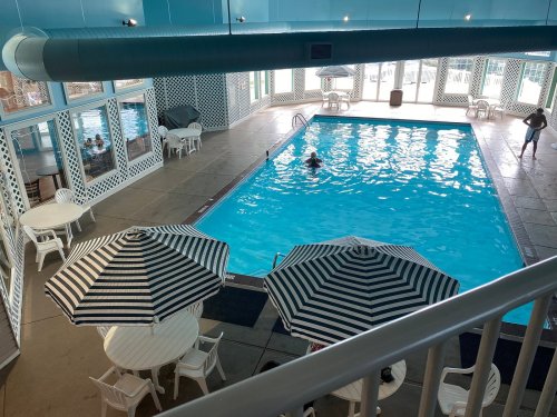 Indoor swimming pool with striped umbrellas, tables, and people enjoying the water. Bright and spacious atmosphere.