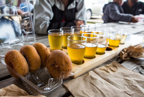 A wooden tray holds small glasses of golden liquid alongside a tray of cinnamon sugar donuts on a picnic table.