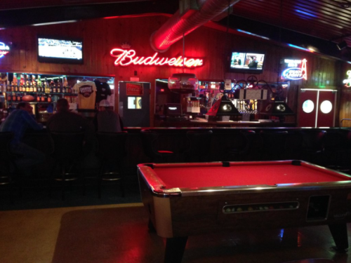 A dimly lit bar with a pool table in the foreground and a neon Budweiser sign, featuring a long bar and TVs.