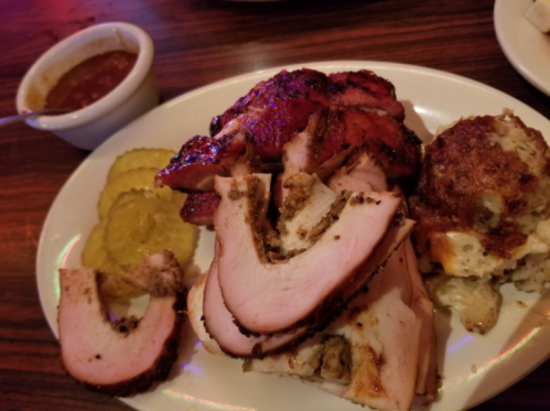 A plate of sliced smoked meat, pickles, and a side of cornbread with sauce in a small bowl.