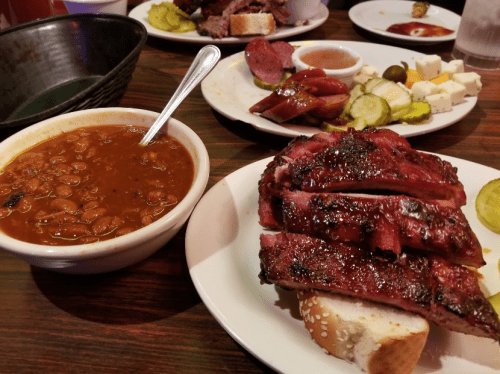 A plate with sliced barbecue ribs on bread, a bowl of beans, and a platter of meats and pickles in the background.