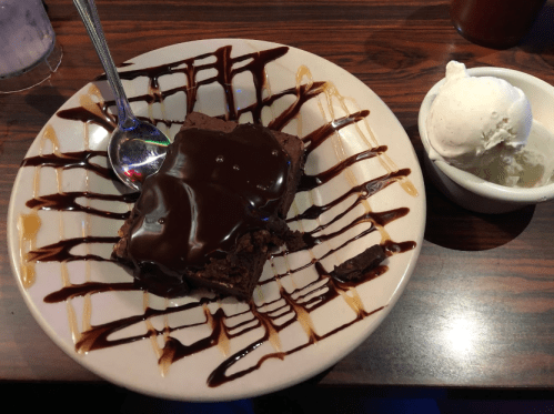 A plate with a chocolate brownie drizzled in sauce, accompanied by a scoop of vanilla ice cream in a small bowl.