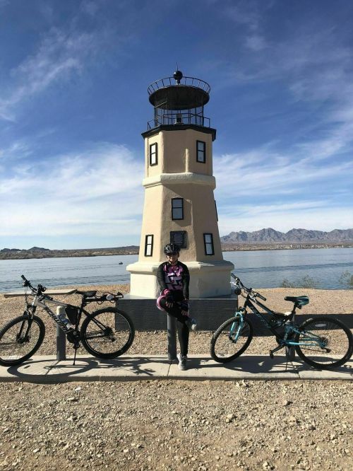 A person in cycling gear stands between two bicycles near a lighthouse by a lake, with mountains in the background.