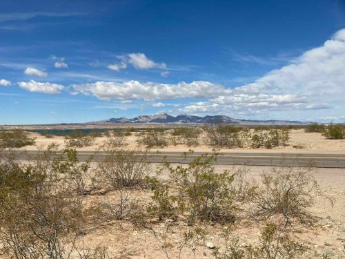 A desert landscape with sparse vegetation, mountains in the distance, and a blue sky with scattered clouds.