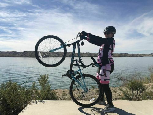 A person in cycling gear lifts a mountain bike above their head by the front wheel near a calm lake.