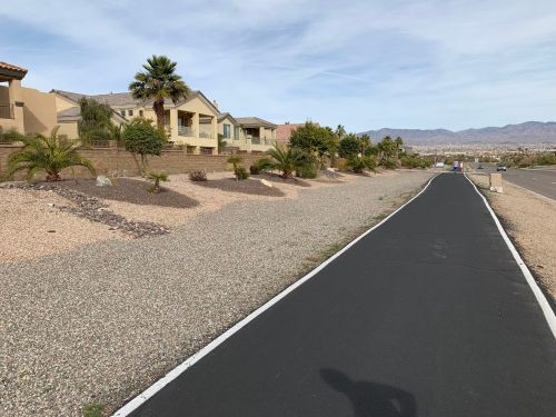 A paved path alongside a desert landscape with houses and palm trees in the background under a clear sky.