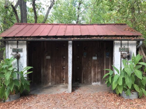 Two rustic wooden restroom doors labeled "GENTS" and "LADIES," surrounded by greenery and a red metal roof.