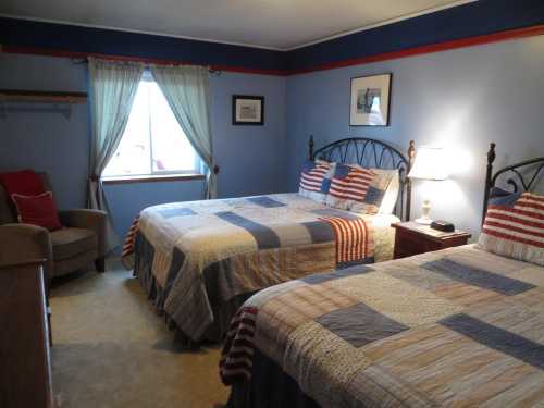 A cozy bedroom with two beds, striped quilts, a window with curtains, and a chair in the corner.