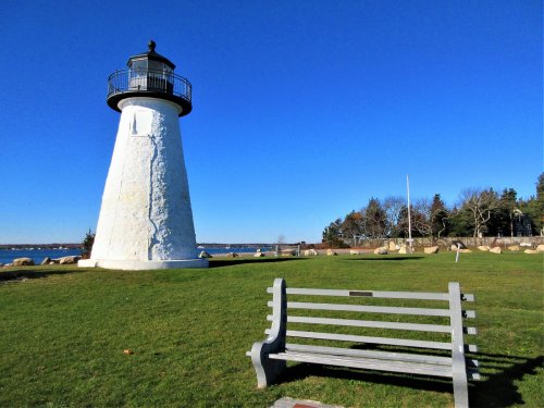 A white lighthouse stands beside a grassy area with a bench, under a clear blue sky and near the water.