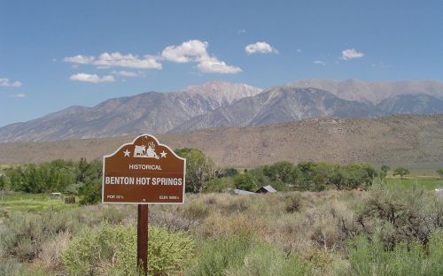 Sign for Benton Hot Springs with mountains and blue sky in the background, surrounded by shrubs and greenery.