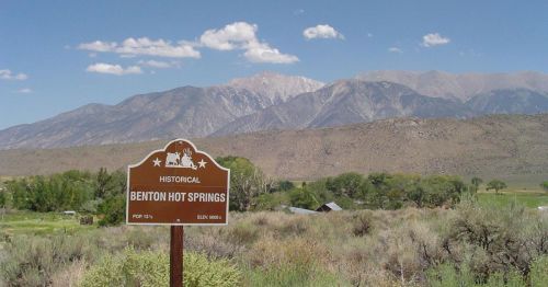 Sign for Benton Hot Springs in front of mountains and a clear blue sky, surrounded by shrubs and grass.
