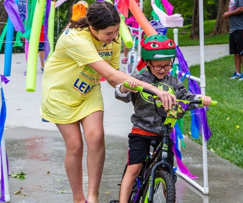 A girl helps a boy in a helmet ride a bike through a colorful, festive obstacle course with streamers and pool noodles.