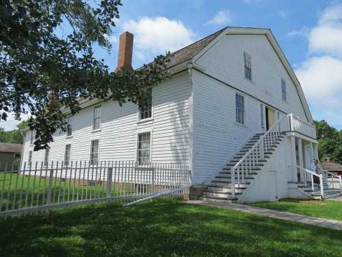 A white two-story house with a sloped roof, stairs, and a picket fence, surrounded by green grass and blue sky.