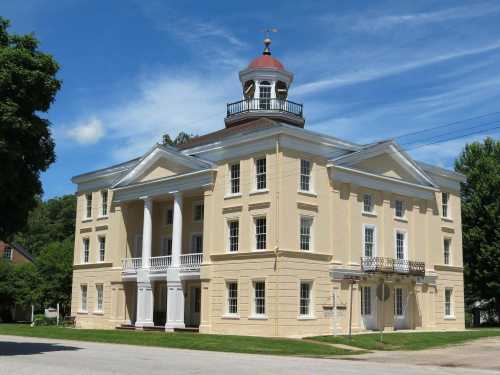 Historic yellow building with a red dome, white columns, and a green lawn under a blue sky.