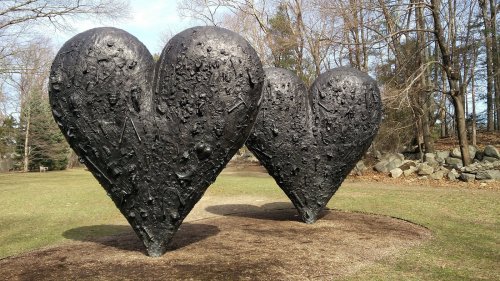 Two large, black heart-shaped sculptures in a grassy area surrounded by trees and rocks.