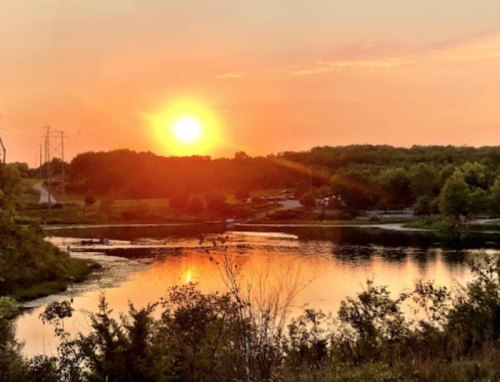 Sunset over a tranquil lake, with trees and a road in the background, reflecting warm colors on the water's surface.
