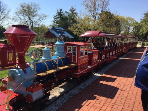 A colorful miniature train with a red engine and blue cars, parked on a brick path in a park setting.