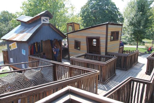 Two colorful playhouses surrounded by wooden decks and railings in a park setting with trees in the background.