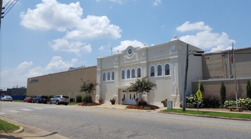 A white building with decorative architecture, flanked by a larger structure, under a blue sky with fluffy clouds.