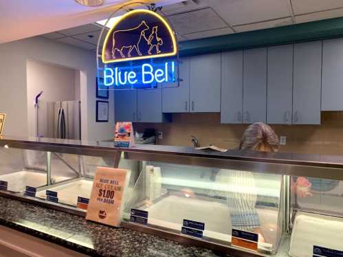 A brightly lit ice cream counter with a "Blue Bell" neon sign and empty ice cream tubs.