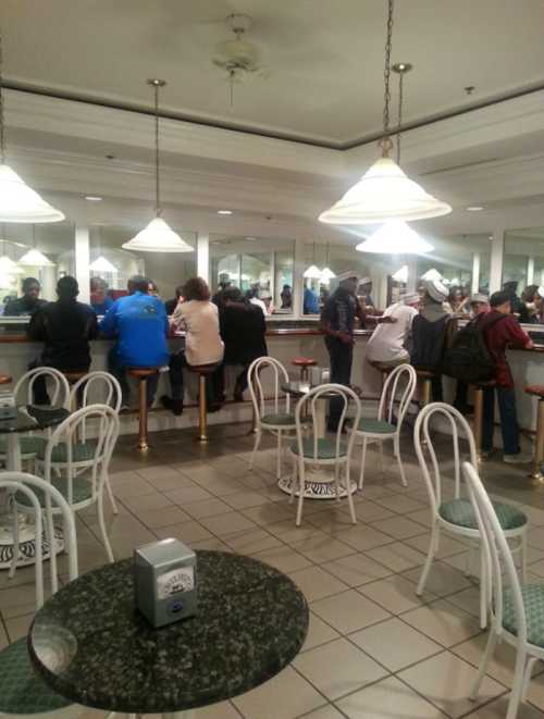 A busy café interior with patrons at the counter and tables, featuring white chairs and pendant lights.