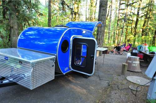 A blue teardrop trailer parked in a forest campsite, with people gathered around a fire pit in the background.