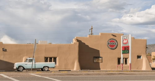A vintage light blue truck parked in front of a southwestern-style building with a Route 66 sign and gas prices.