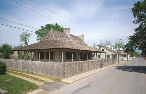 A historic house with a thatched roof, surrounded by a wooden fence, on a quiet street lined with trees.
