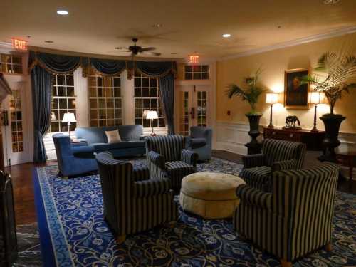 Cozy lounge area with blue sofas, striped chairs, a round ottoman, and large windows, decorated with plants and artwork.