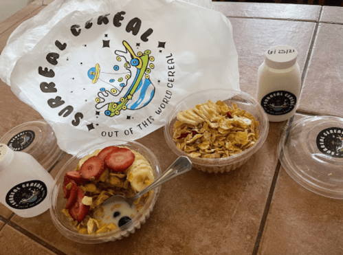 Two bowls of cereal topped with banana and strawberry slices, a plastic bag labeled "Surreal Cereal," and milk bottles.