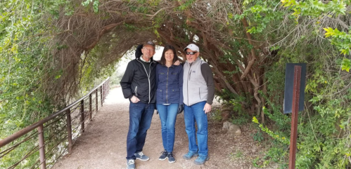 Three people stand together under a leafy archway on a nature path, smiling at the camera.