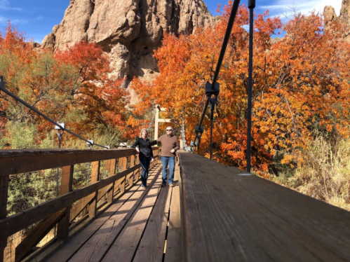 Two people walk hand in hand across a wooden bridge surrounded by vibrant autumn foliage.