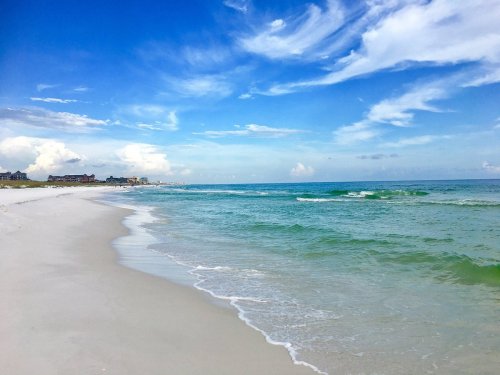 A serene beach scene with soft white sand, gentle waves, and a bright blue sky with scattered clouds.