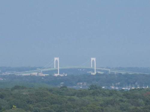 A distant view of a suspension bridge spanning a river, surrounded by green trees and a hazy sky.