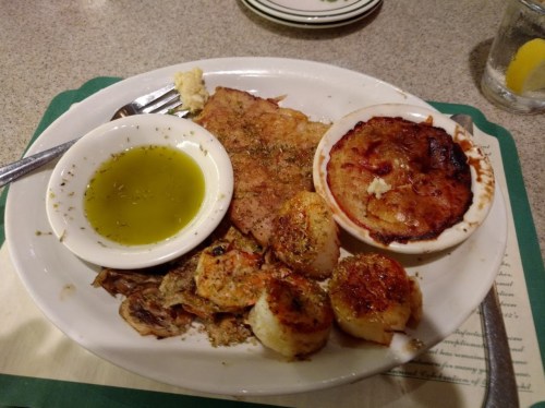 A plate of seafood featuring fried fish, scallops, and a baked dish, served with olive oil and herbs on the side.