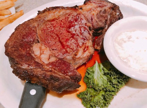 A large, juicy ribeye steak on a plate, accompanied by a side of creamy dip and a garnish of kale.