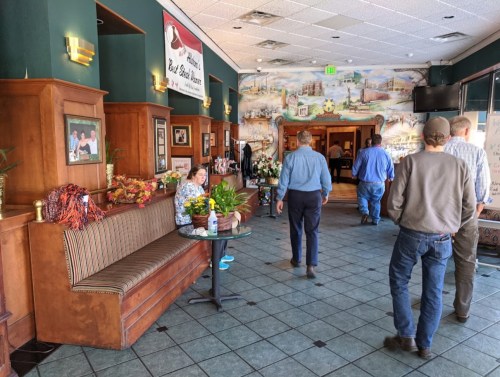 A restaurant interior with wooden decor, a woman sitting on a bench, and patrons walking towards the entrance.