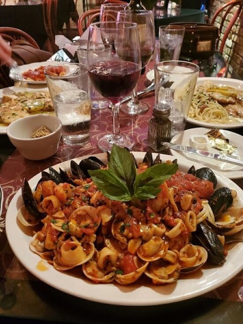 A plate of seafood pasta with clams and mussels, surrounded by glasses of wine and other dishes on a table.