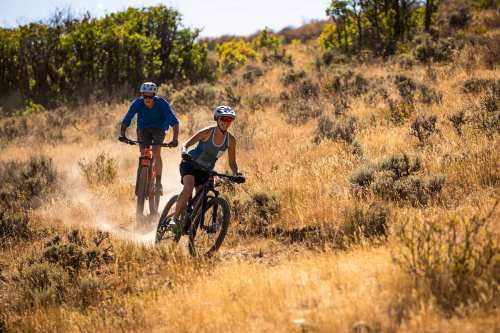 Two mountain bikers ride on a dusty trail through a grassy landscape with shrubs and trees in the background.