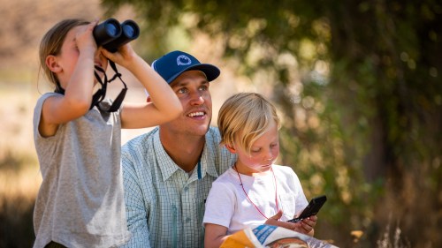 A man smiles while two children explore nature, one using binoculars and the other looking at a phone.