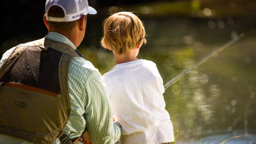 A person and a child fishing together by a serene water body, enjoying a sunny day outdoors.