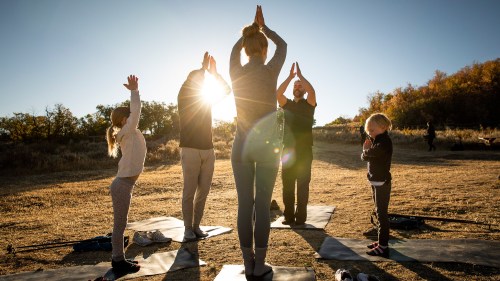 A group of people practicing yoga outdoors at sunset, with hands raised in a prayer position.