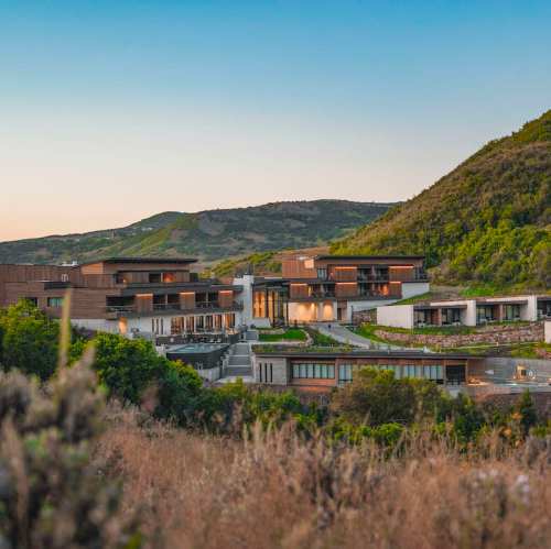 Modern buildings nestled in a hillside landscape, surrounded by greenery and mountains under a clear sky.