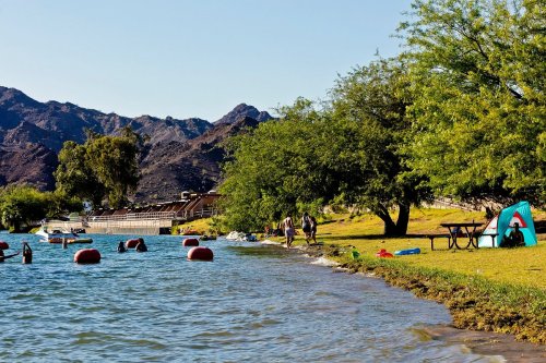 A serene lakeside scene with people swimming, a tent, and mountains in the background under a clear blue sky.