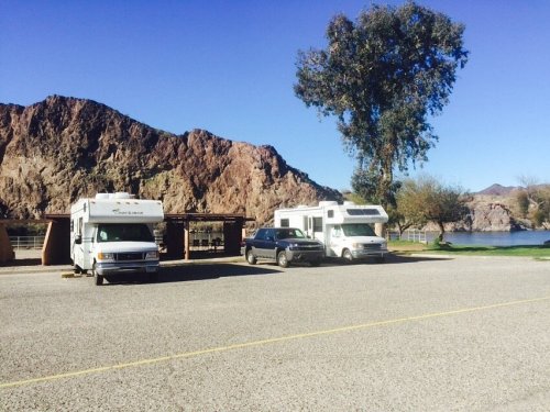 Two RVs and a car parked near a lake, with rocky mountains and trees in the background under a clear blue sky.