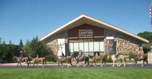 A line of horseback riders passes by the Buffalo Bill Center of the West under a clear blue sky.